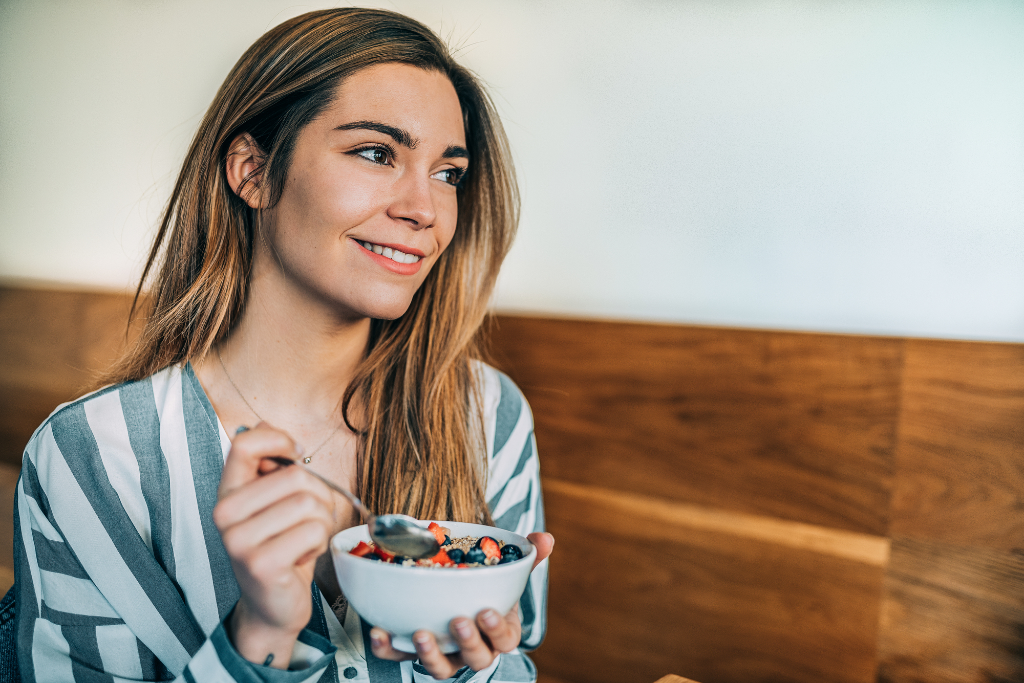 A woman with long hair smiles while holding a bowl of food topped with blueberries and strawberries. She is wearing a striped shirt and sitting in a room with wooden paneling on the walls. She holds a spoon in her other hand as she looks to the side.