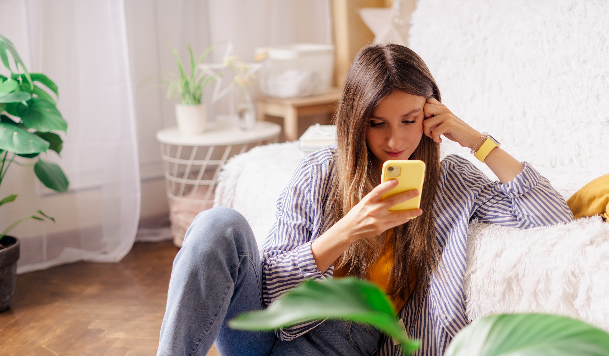 A woman with long hair, dressed in a striped shirt and jeans, is sitting on the floor and leaning against a white couch. She is looking at her yellow smartphone, with one hand resting on her forehead. There are houseplants and soft furnishings in the cozy, bright room.