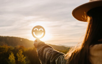 A person holds up a circular cutout with a smiling face, positioned to catch the sunlight, creating a glowing effect. The person is wearing a hat and flannel shirt, standing in a scenic outdoor area with trees and rolling hills in the background.