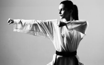 Black and white photo of a person in a martial arts uniform practicing a punch. They have a determined expression and are positioned sideways, extending one arm straight while keeping the other arm bent. They are wearing a black belt and have their hair tied back.