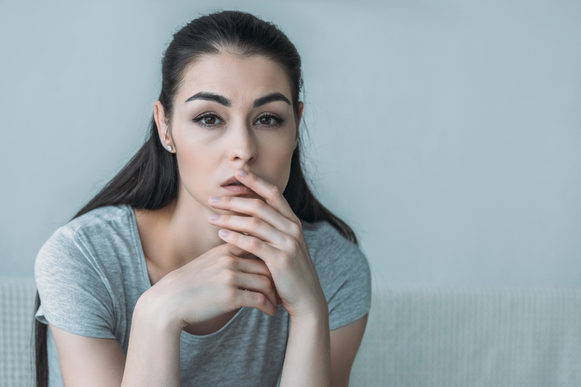 A young woman with long dark hair and wearing a grey shirt is sitting indoors. She has a thoughtful expression, resting her chin on her hand. The background is blurred, focusing attention on her face.