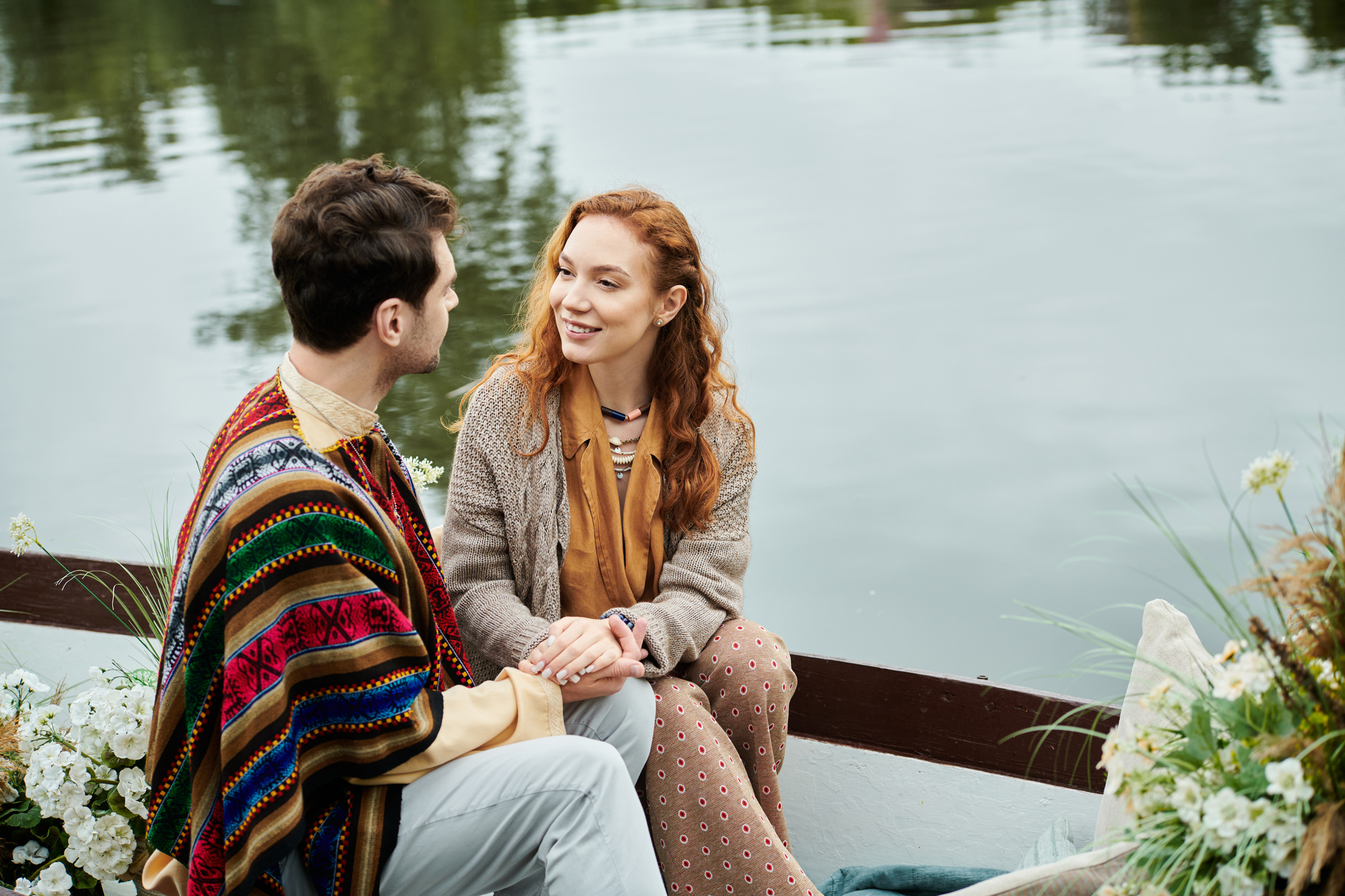 A couple sits in a boat on a calm lake, surrounded by flowers and greenery. The man is wearing a colorful poncho, and the woman has curly red hair and is smiling while holding his hand. They are engaging in a warm conversation.