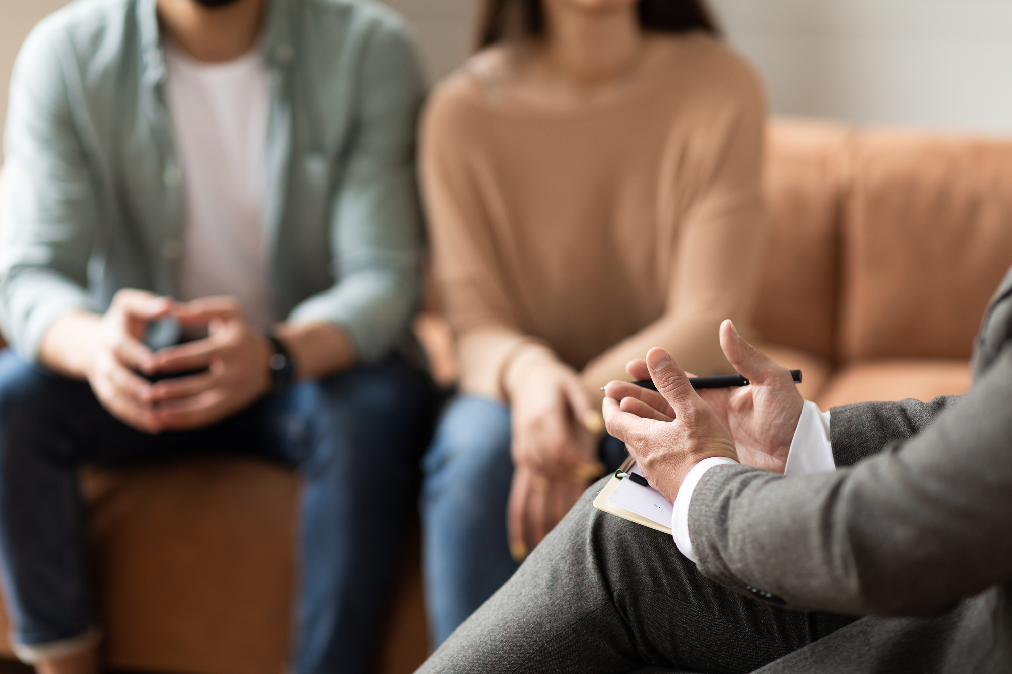 A close-up of a therapist's hands holding a pen and notepad, gesturing while speaking. In the background, a couple sits on a couch, attentively listening with their hands placed on their laps. The setting appears to be a counseling or therapy session.