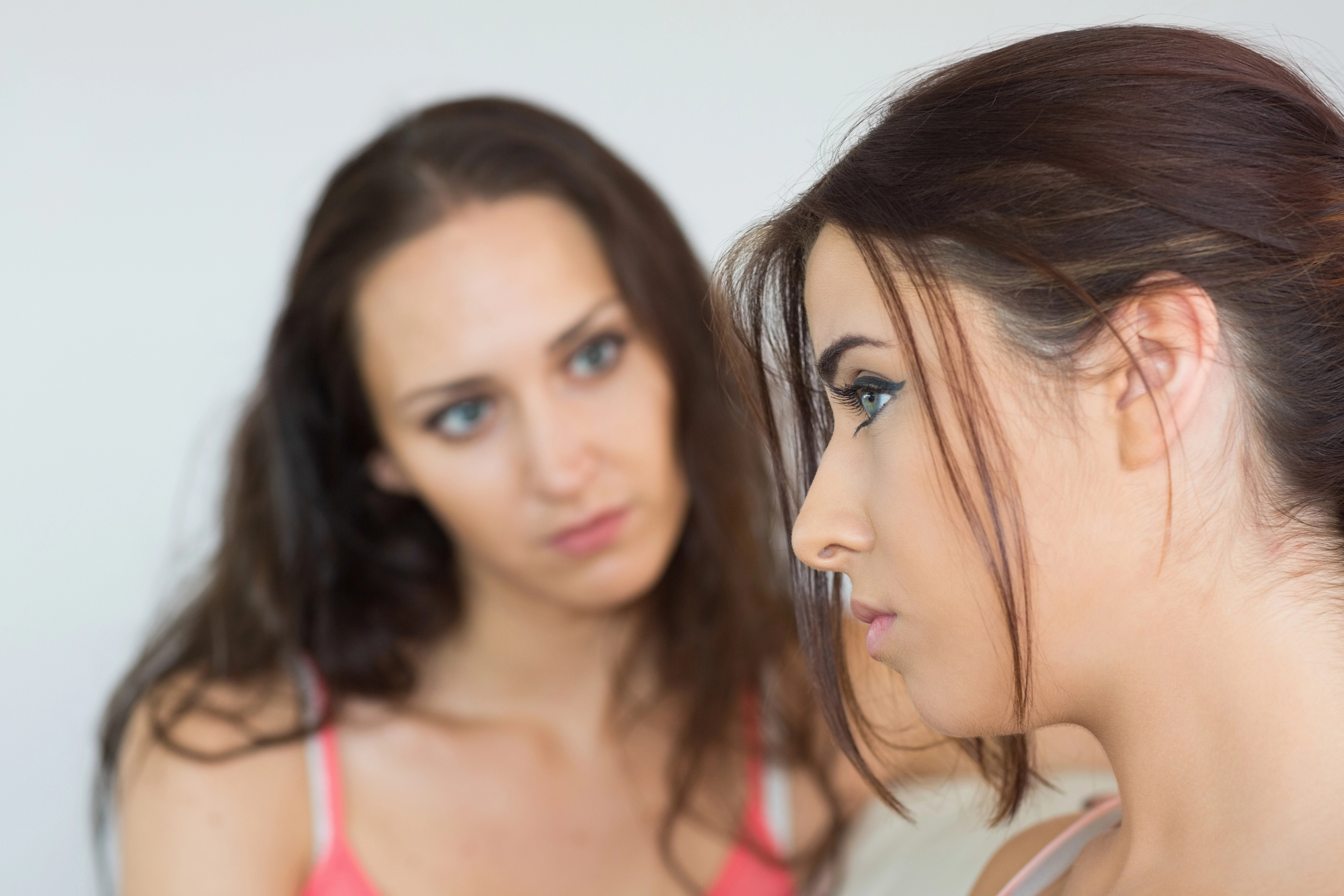 Two women are in the image, one in the foreground and the other in the background. The woman in the foreground has brown hair, tied back, and is looking down with a serious expression. The woman in the background has long brown hair and is also looking serious.