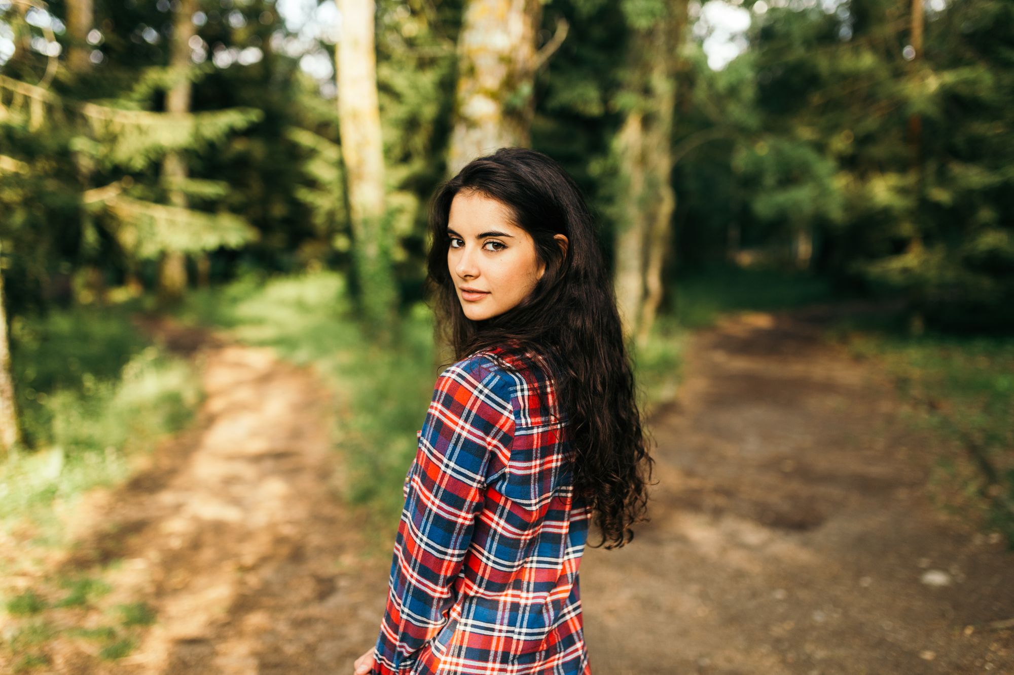 A woman with long, dark hair stands on a forest path, wearing a red, blue, and white plaid shirt. She is looking over her shoulder toward the camera with trees and greenery surrounding her.