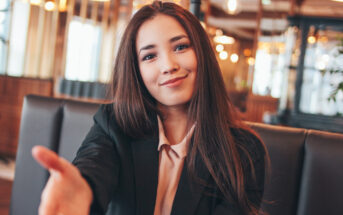 A young woman with long brown hair wearing a black blazer and light pink blouse sits in a warmly lit cafe, extending her hand forward with a welcoming smile. The background features modern decor with hanging lights, wooden accents, and large windows.