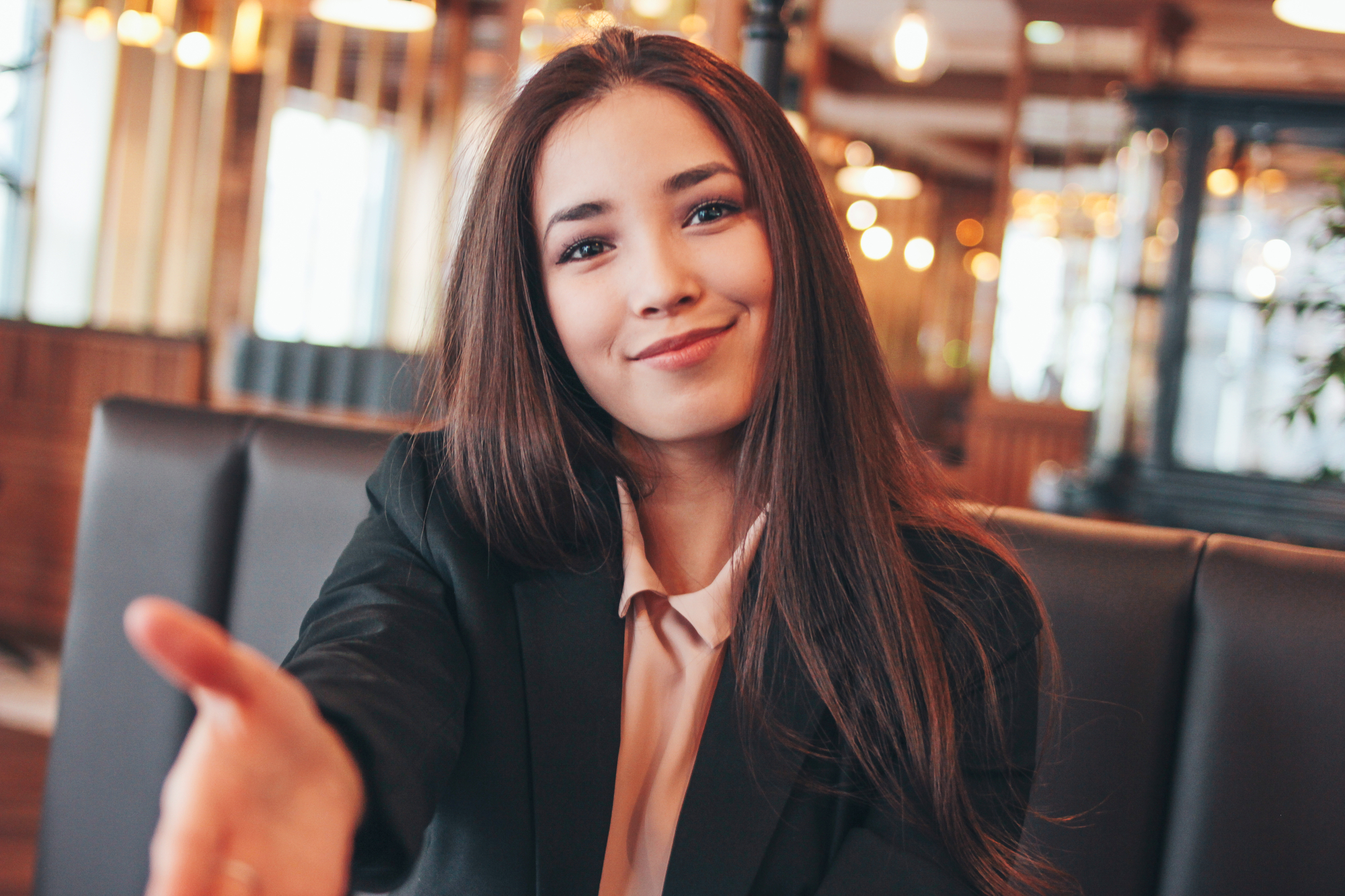 A young woman with long brown hair wearing a black blazer and light pink blouse sits in a warmly lit cafe, extending her hand forward with a welcoming smile. The background features modern decor with hanging lights, wooden accents, and large windows.