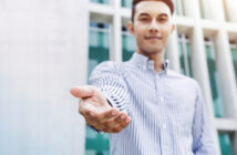 A person wearing a striped button-up shirt extends their hand forward in a welcoming gesture. The background features a modern building with a grid-like glass facade. The image is slightly blurred, focusing on the gesture.