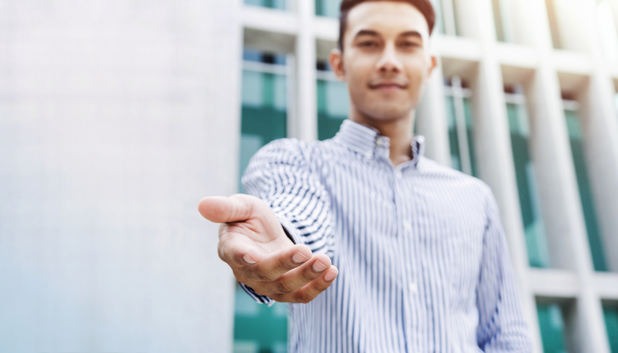 A person wearing a striped button-up shirt extends their hand forward in a welcoming gesture. The background features a modern building with a grid-like glass facade. The image is slightly blurred, focusing on the gesture.