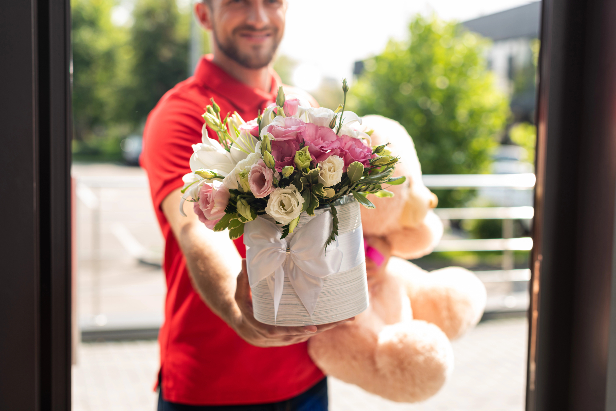 A person in a red shirt is holding a bouquet of flowers and a large teddy bear, presenting them towards the viewer as if making a delivery. The background shows a bright, outdoor setting.