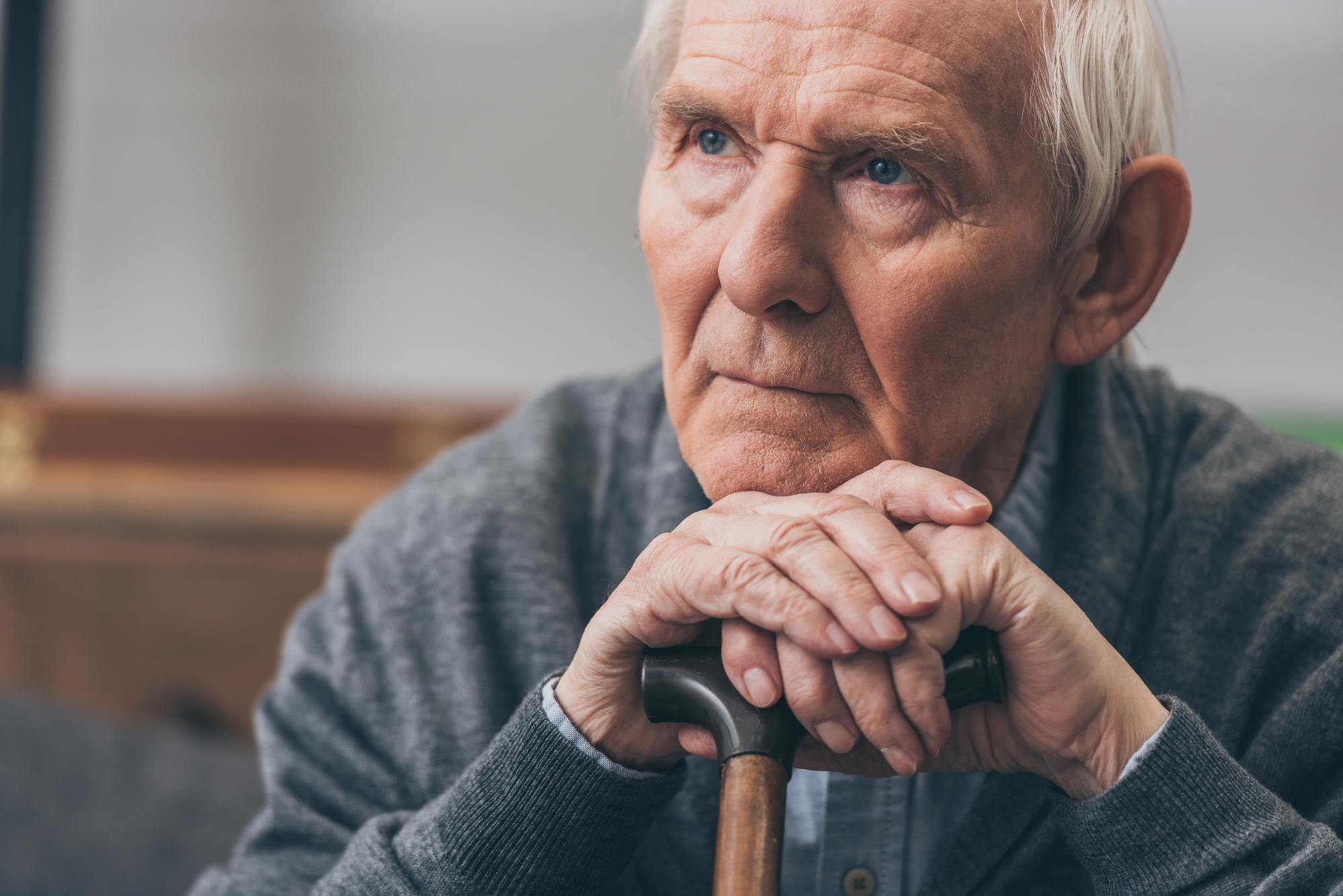 An elderly man with white hair rests his chin on his hands, which are placed on top of a walking cane. He is wearing a grey sweater and appears deep in thought, looking slightly upwards. The background is out of focus.