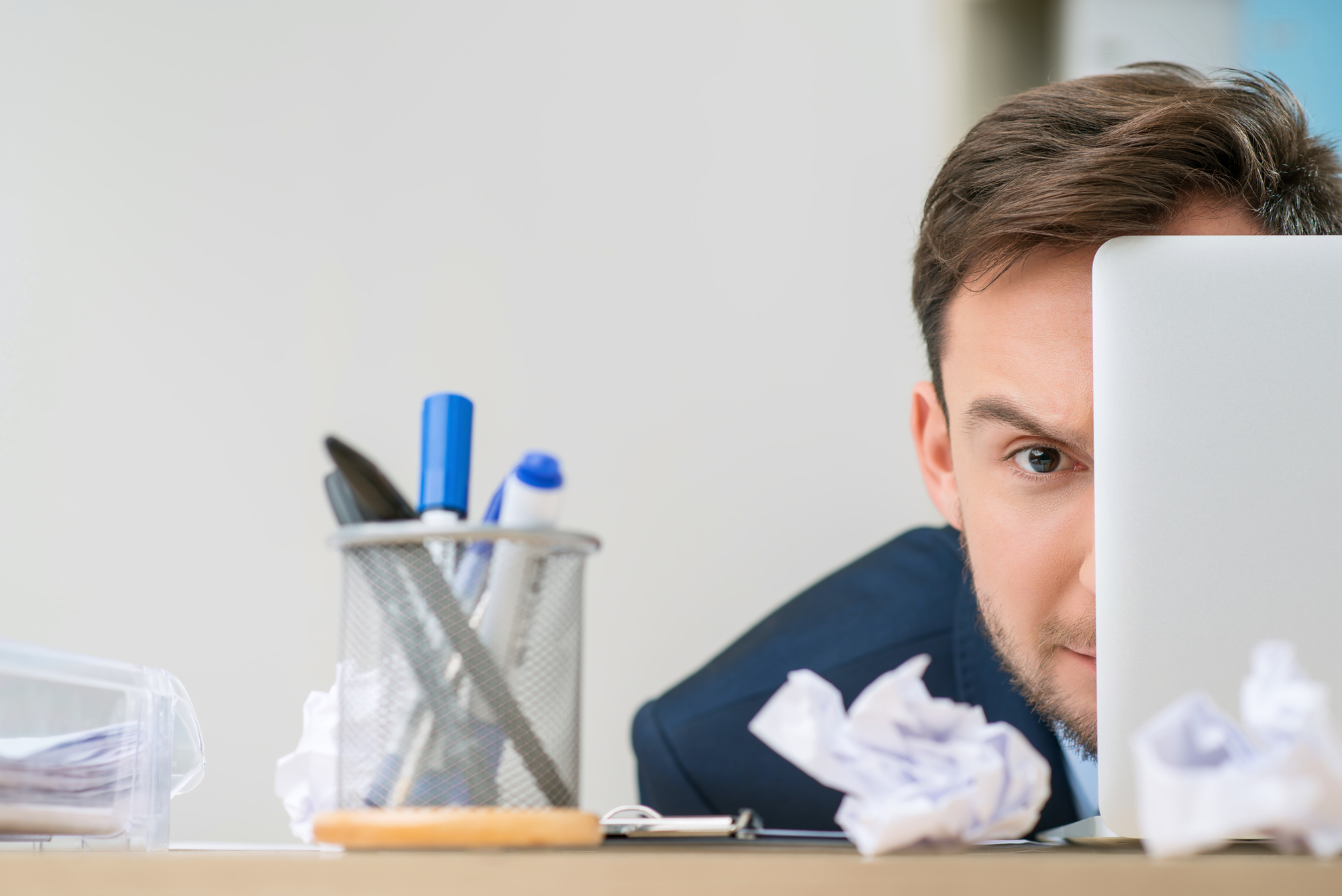 A man in a suit is partially hidden behind a laptop screen. He is looking intently at the camera. The desk in front of him is cluttered with crumpled paper, a pen holder containing pens and scissors, and other office supplies.