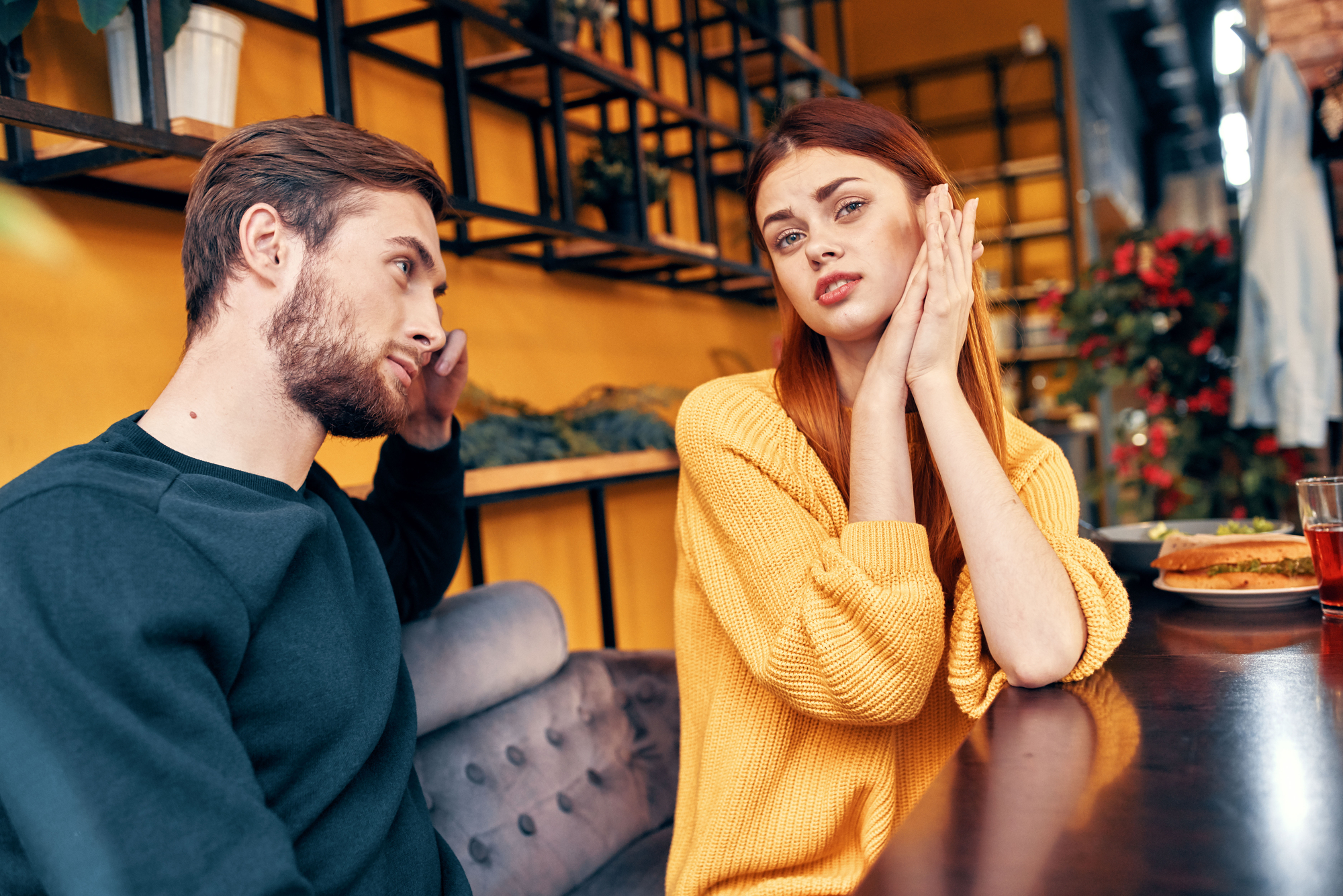 A man and a woman are sitting in a cafe, engaged in a conversation. The woman, wearing a yellow sweater, appears distressed or frustrated, with her hands raised to her face. The man, in a dark sweater, looks at her attentively. The background is decorated with shelves and plants.