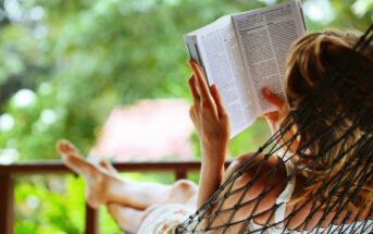 A person with blond hair is lying on a hammock outdoors, reading a book. The scene is set in a lush, green environment with a blurred background of trees and foliage. The person's feet are propped up, adding to the relaxed ambiance.