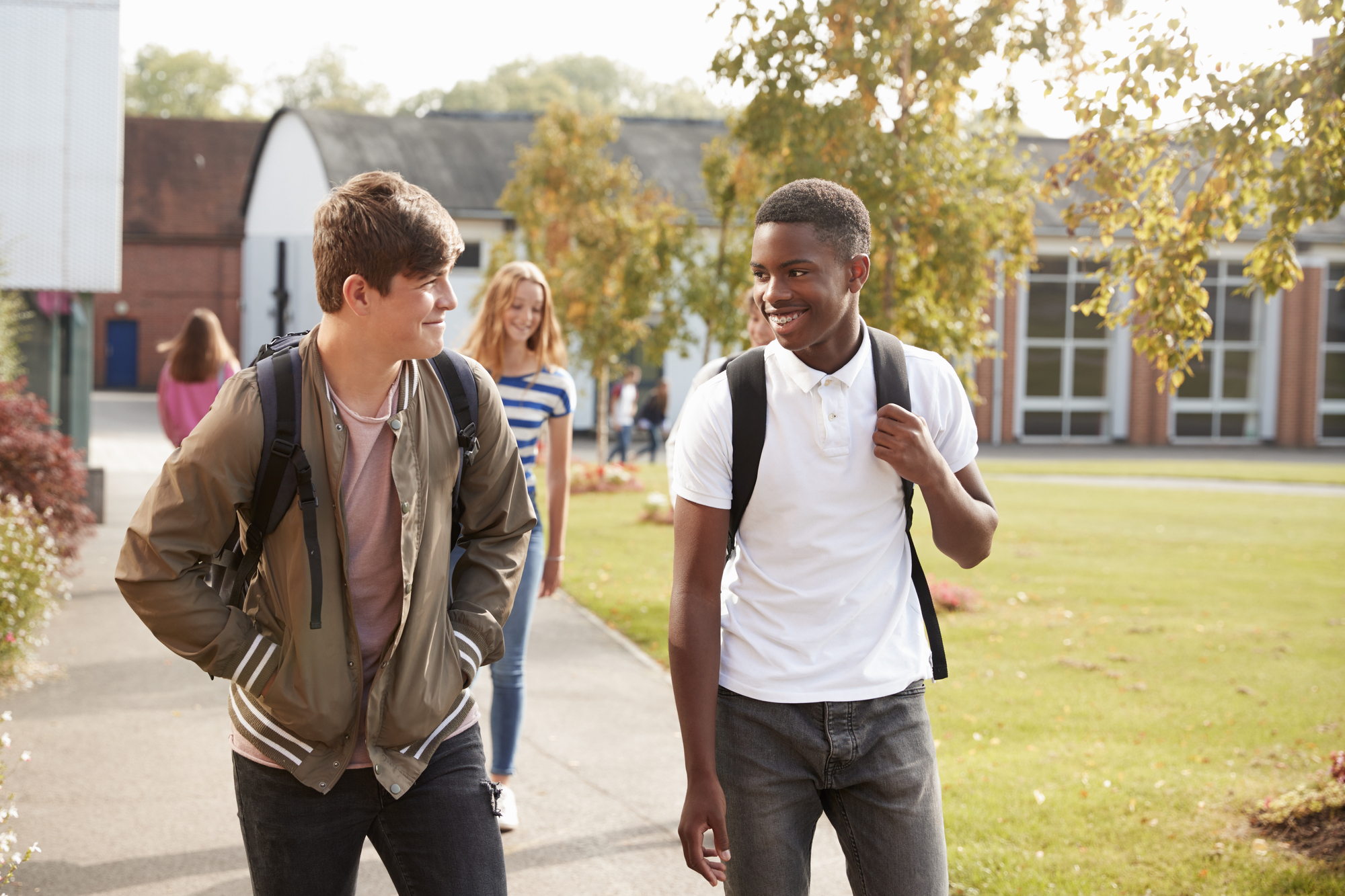 Two young men, one in a brown jacket and the other in a white polo, walk together and smile on a sunny school campus. They each have backpacks. A girl in a blue and white striped shirt walks behind them near trees and a brick building.