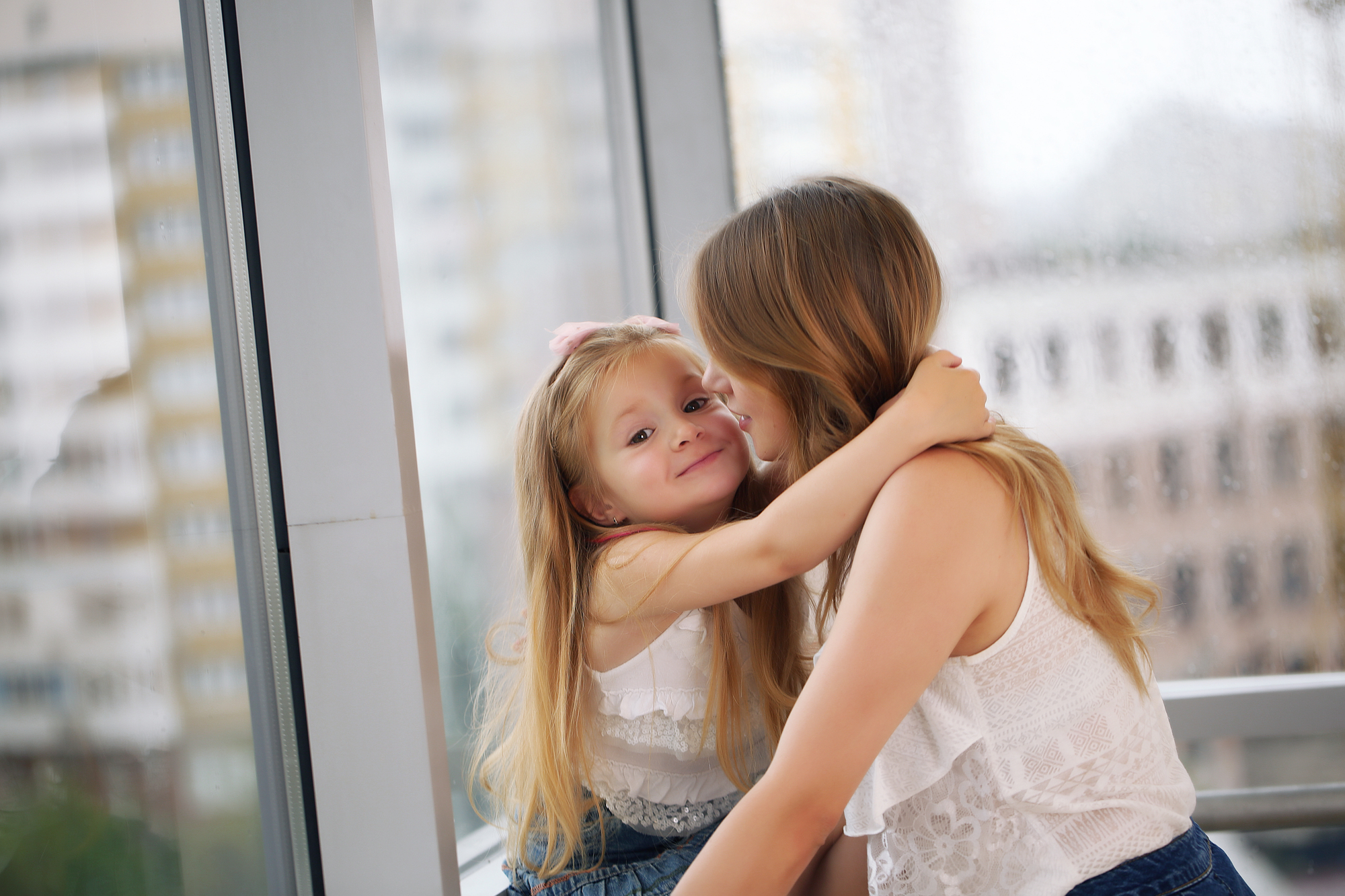 A woman and a young girl with long blonde hair are sitting by a large window. The girl is embracing the woman, who is smiling and pressing their cheeks together. Both are wearing white tops and jeans. Outside the window, a blurred cityscape is visible.