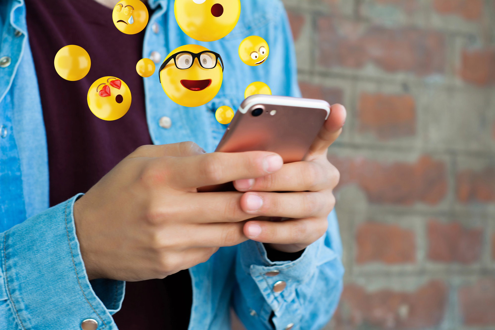 A person wearing a denim jacket is holding a smartphone while various emojis, including a heart-eyed emoji, a glasses-wearing smiley, and others, appear to float around the phone screen against a brick wall background.