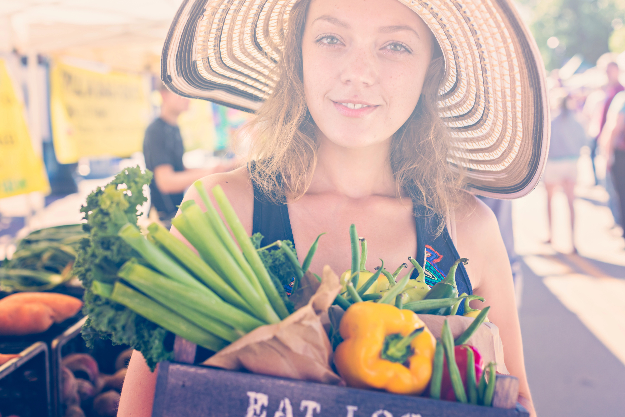 A woman wearing a large sun hat smiles while holding a wooden crate filled with fresh vegetables, including celery stalks, green beans, and a yellow bell pepper. The background shows an outdoor market with other people and vendor stalls.