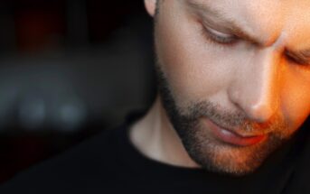 Close-up of a man with a trimmed beard and short hair looking down with a serious expression. The lighting casts a warm glow on the right side of his face while the background is blurred and dark. He is wearing a black shirt.