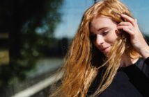A person with long, red hair stands outdoors, lightly touching their hair with one hand. They are wearing a black long-sleeved top and appear to be in bright sunlight. The background is slightly blurred, showing trees and other natural elements.