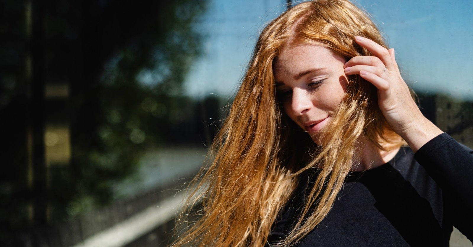 A person with long, red hair stands outdoors, lightly touching their hair with one hand. They are wearing a black long-sleeved top and appear to be in bright sunlight. The background is slightly blurred, showing trees and other natural elements.