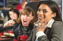 A woman in a business suit smiles and looks at the camera while sitting in a cafe. Behind her, another woman with brown hair is angrily yelling and gesturing with her fists, and a third woman watches. The scene seems to indicate conflict or aggression.