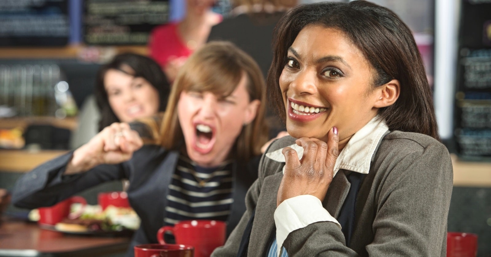 A woman in a business suit smiles and looks at the camera while sitting in a cafe. Behind her, another woman with brown hair is angrily yelling and gesturing with her fists, and a third woman watches. The scene seems to indicate conflict or aggression.