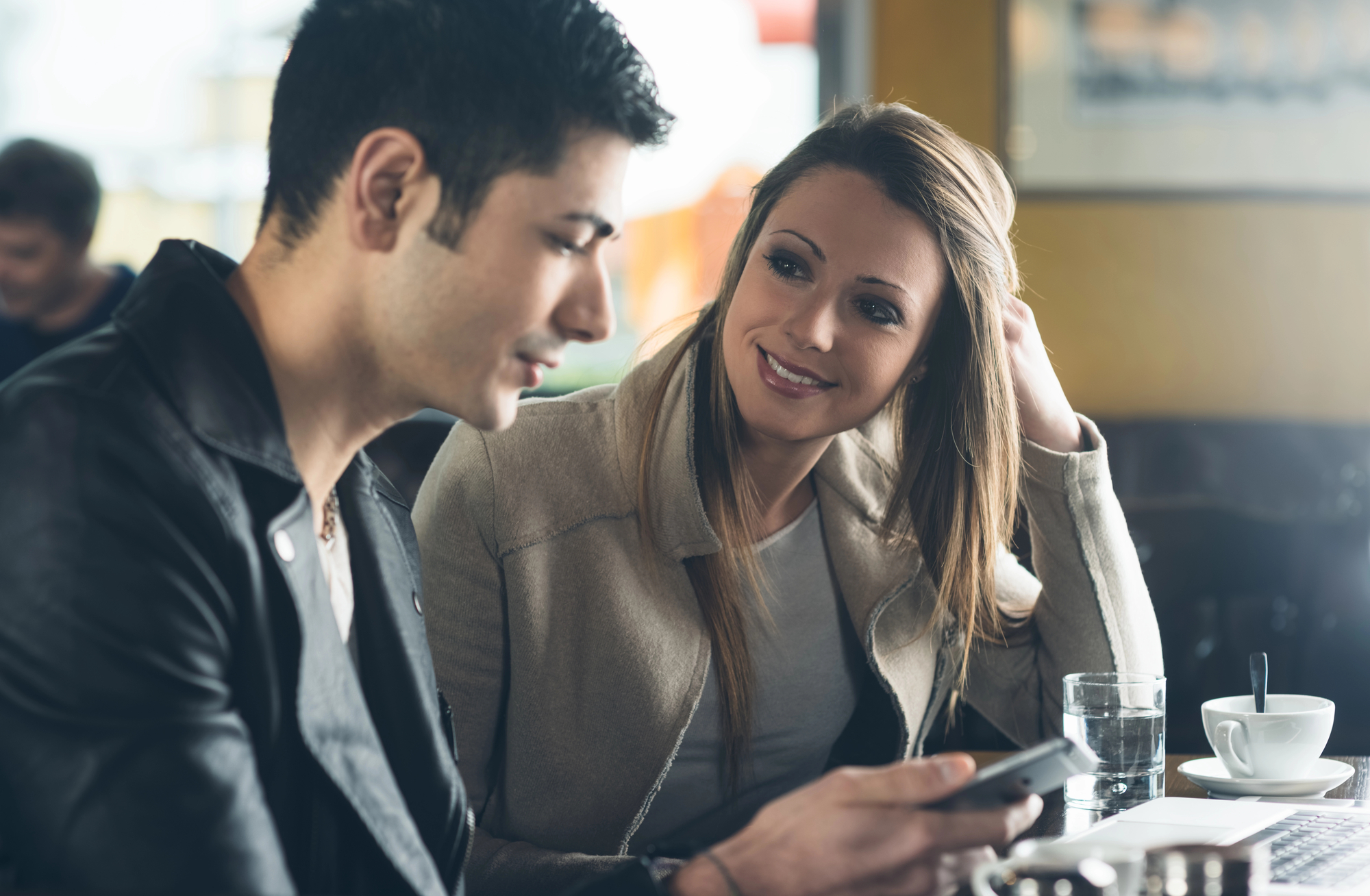 A man and woman sit at a cafe table, the man looking at his smartphone while the woman smiles at him. The woman has long hair and is wearing a beige coat, and the man is wearing a black leather jacket. There are cups on the table.