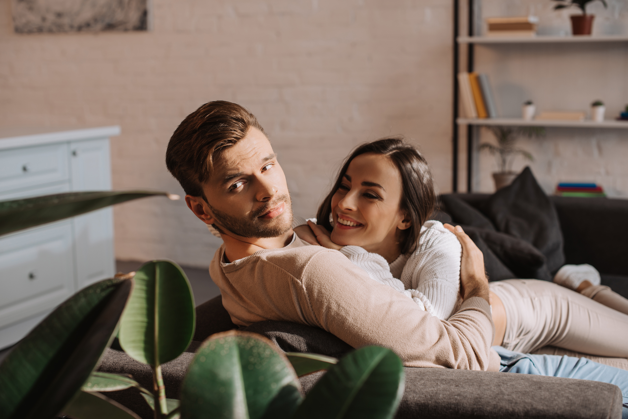 A couple is sitting on a couch in a cozy, modern living room. The man, with short brown hair and a beard, looks contemplatively at the camera, while the woman, with long dark hair, smiles and wraps her arms around him. There are books and plants in the background.