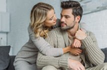 A woman with blonde hair, wearing a gray sweater, embraces a man with brown hair, in a beige shirt and knitted cardigan. They sit closely on a couch, smiling and looking into each other's eyes, conveying affection and warmth.