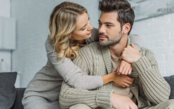 A woman with blonde hair, wearing a gray sweater, embraces a man with brown hair, in a beige shirt and knitted cardigan. They sit closely on a couch, smiling and looking into each other's eyes, conveying affection and warmth.