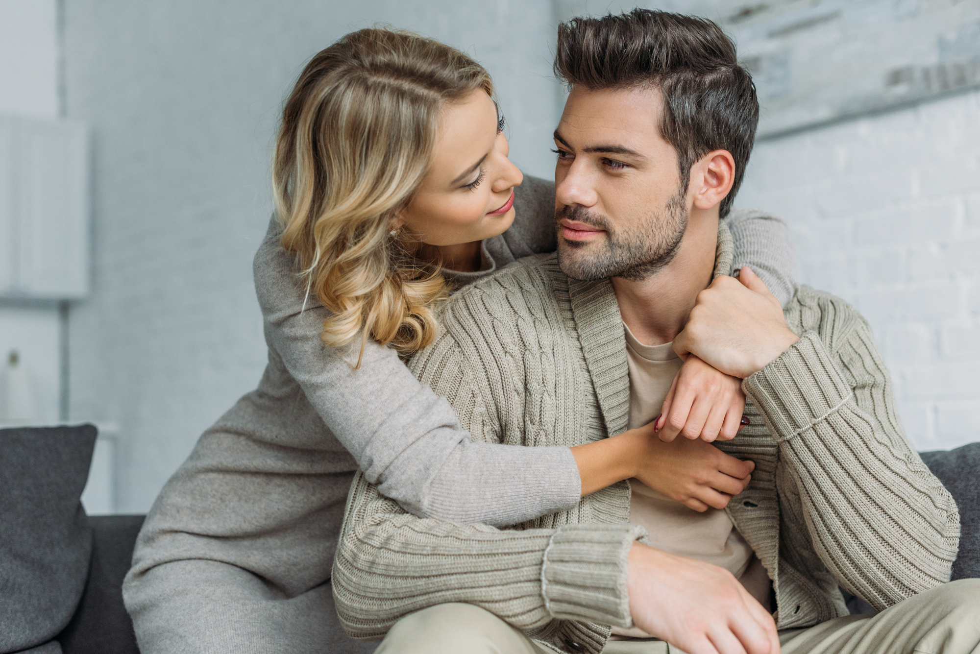 A woman with blonde hair, wearing a gray sweater, embraces a man with brown hair, in a beige shirt and knitted cardigan. They sit closely on a couch, smiling and looking into each other's eyes, conveying affection and warmth.