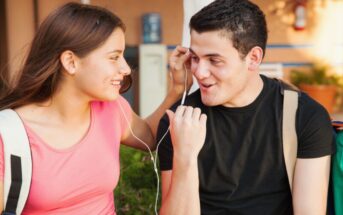 Two young people are listening to music together, sharing a pair of earphones. They are smiling and appear to be enjoying each other's company. Both carry backpacks, suggesting they might be students. The background is slightly blurred with a plant and a building visible.