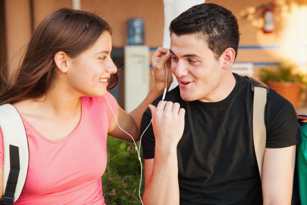 Two young people are listening to music together, sharing a pair of earphones. They are smiling and appear to be enjoying each other's company. Both carry backpacks, suggesting they might be students. The background is slightly blurred with a plant and a building visible.