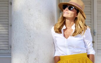 Woman leaning against a pillar wearing a straw hat, sunglasses, white blouse, and yellow skirt. She is looking up and smiling, with white shutters in the background. The scene appears to be in a warm, sunny outdoor setting.
