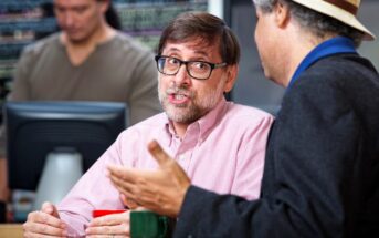 Two men are engaged in a conversation in an office or work environment. One man with glasses and a beard is wearing a pink shirt and holding a red cup, while the other man wears a hat and dark jacket. A third person working is in the background.