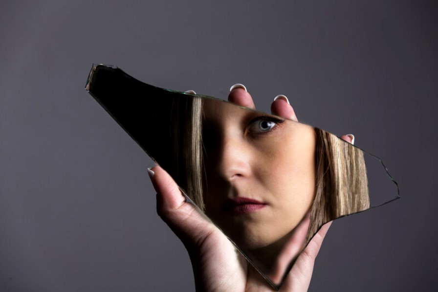 A hand holds a jagged piece of broken mirror. The mirror reveals part of a woman's face, showing her eye and partial features. The background is a plain, dark gray, allowing the reflection to stand out vividly.