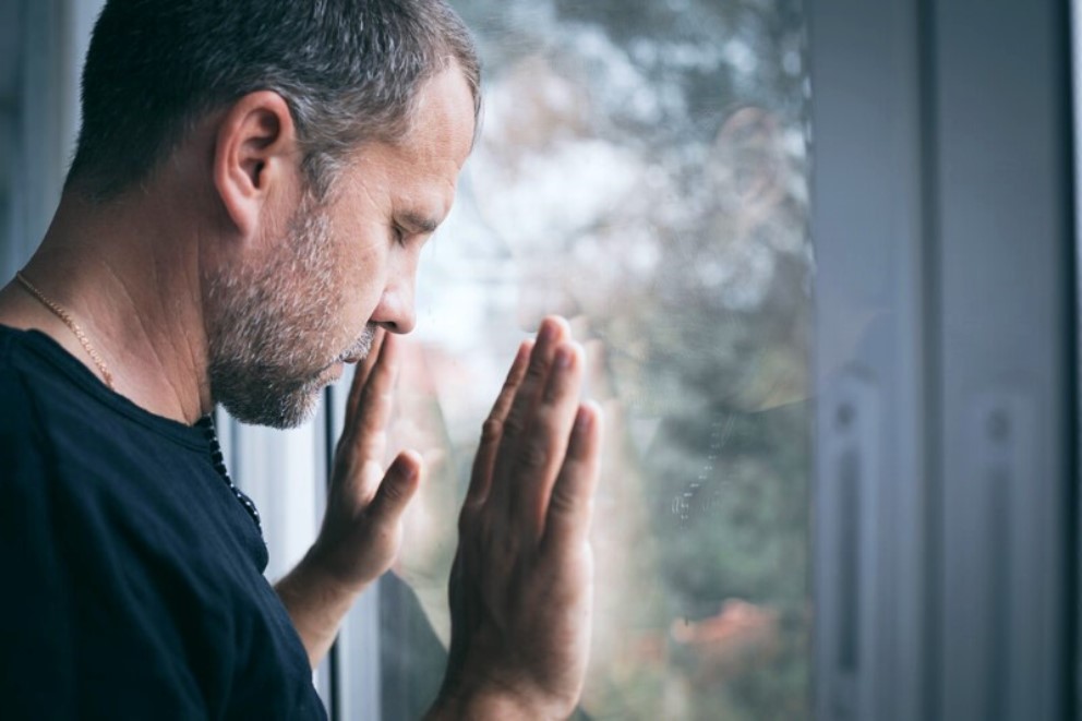 A man with short hair and a beard stands by a window with his hands pressed against the glass. He appears deep in thought or reflection, gazing downwards. The scene outside is blurred, focusing attention on his contemplative expression.