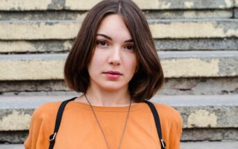 A young woman with shoulder-length brown hair wearing an orange shirt and black suspenders stands in front of a set of weathered concrete steps. She has a calm expression on her face and a long necklace around her neck.