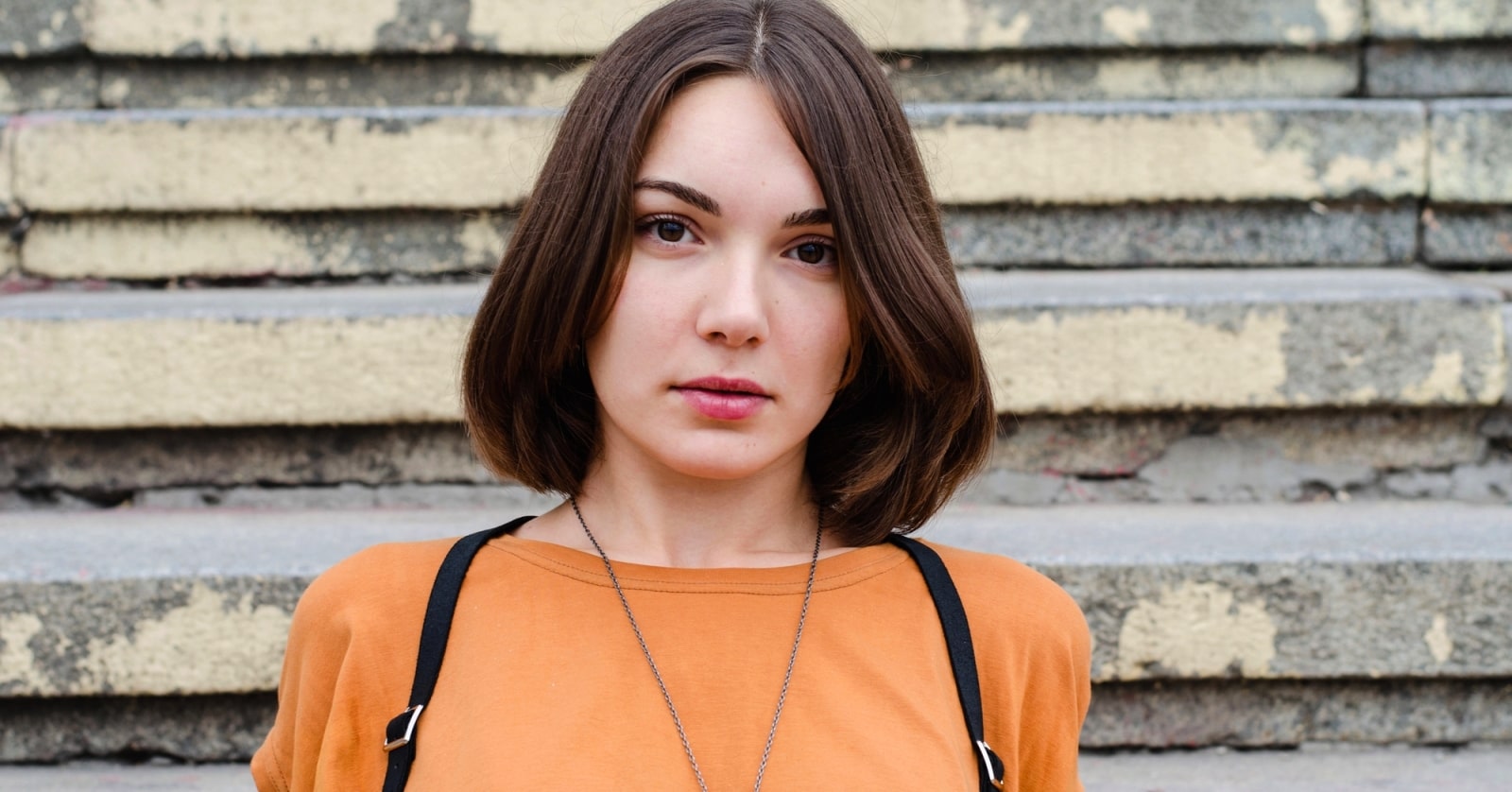 A young woman with shoulder-length brown hair wearing an orange shirt and black suspenders stands in front of a set of weathered concrete steps. She has a calm expression on her face and a long necklace around her neck.