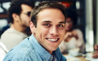 A man with short brown hair, wearing a blue button-down shirt, smiles warmly at the camera. There are people in the background in a softly blurred indoor setting, possibly a cafe.