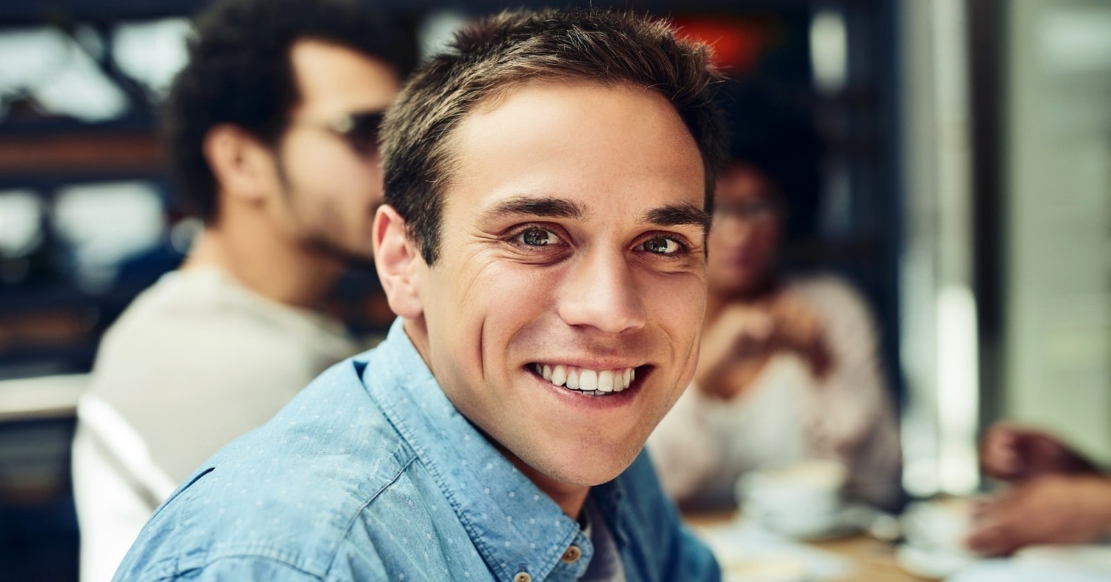 A man with short brown hair, wearing a blue button-down shirt, smiles warmly at the camera. There are people in the background in a softly blurred indoor setting, possibly a cafe.