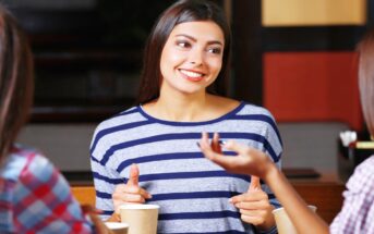 A young woman, smiling and wearing a blue and white striped shirt, chats with two friends over coffee. They are seated in a cozy cafe, and two disposable coffee cups are visible in the foreground.