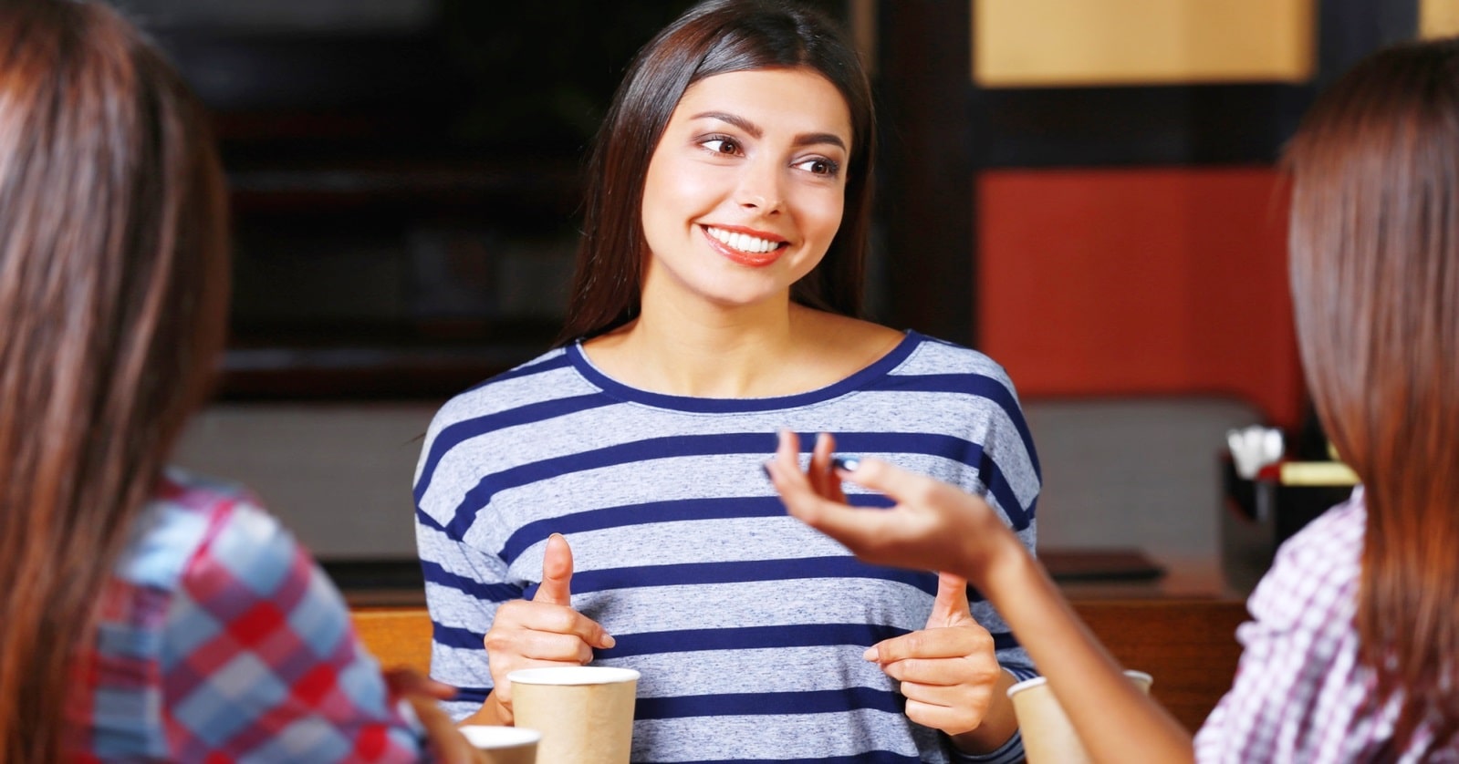 A young woman, smiling and wearing a blue and white striped shirt, chats with two friends over coffee. They are seated in a cozy cafe, and two disposable coffee cups are visible in the foreground.