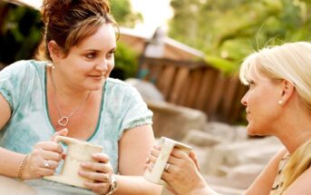 Two women sit outdoors, engaged in a conversation while holding mugs. One woman has brown hair pulled up, and the other has blonde hair. They appear to be sitting in a garden or patio area, with greenery and wooden structures in the background.