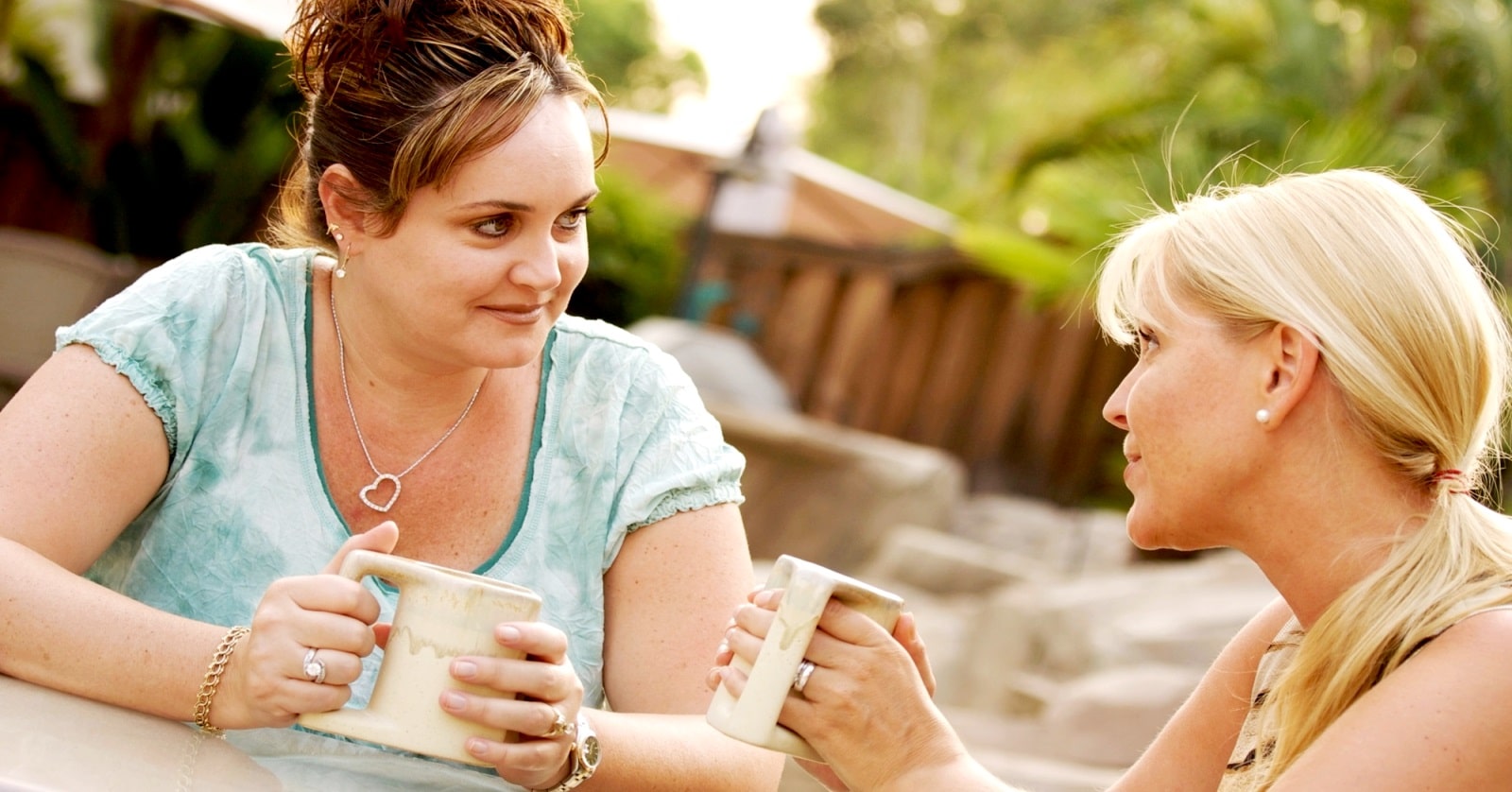 Two women sit outdoors, engaged in a conversation while holding mugs. One woman has brown hair pulled up, and the other has blonde hair. They appear to be sitting in a garden or patio area, with greenery and wooden structures in the background.