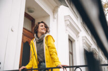A smiling person stands on a balcony, wearing a yellow raincoat and a gray sweater. They are looking off into the distance. The backdrop features a white building with a wooden door and decorative trim.