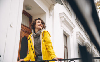 A smiling person stands on a balcony, wearing a yellow raincoat and a gray sweater. They are looking off into the distance. The backdrop features a white building with a wooden door and decorative trim.