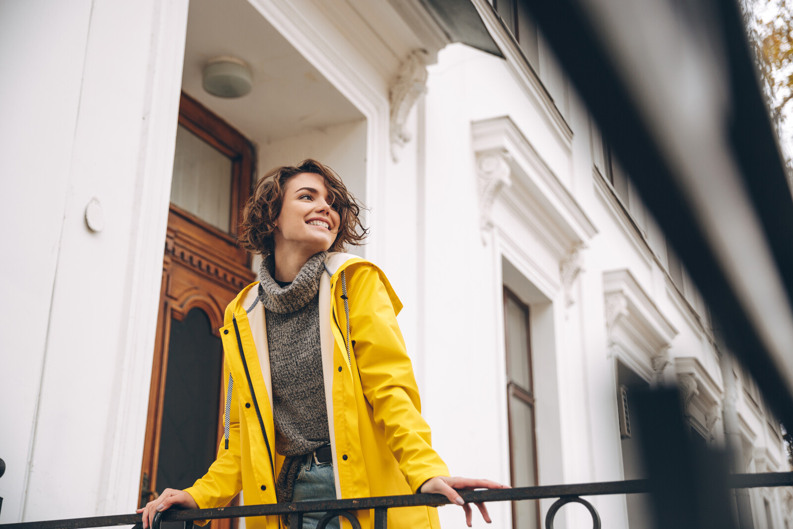 A smiling person stands on a balcony, wearing a yellow raincoat and a gray sweater. They are looking off into the distance. The backdrop features a white building with a wooden door and decorative trim.