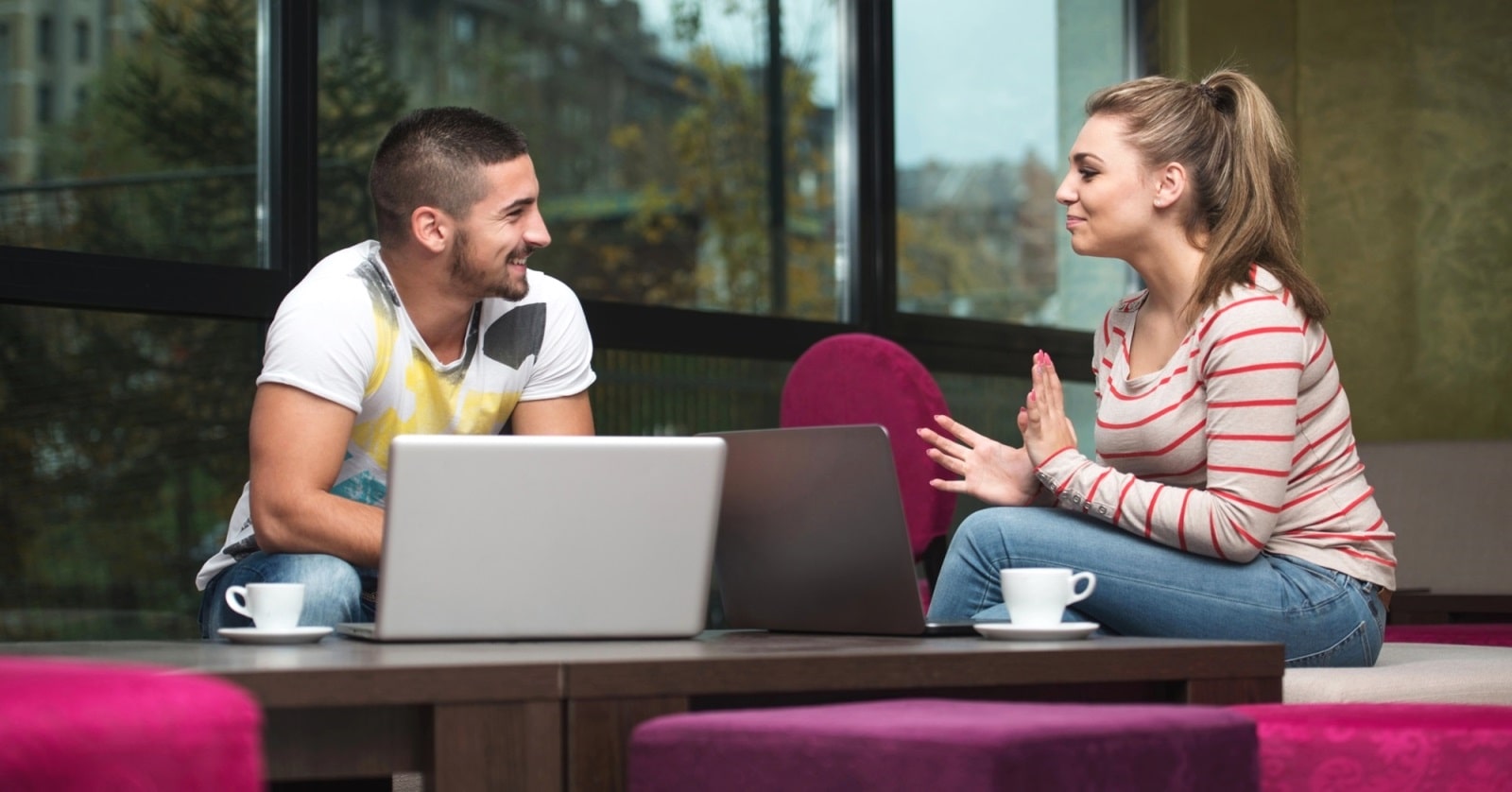 A man and a woman are sitting across a table from each other in a modern, cozy cafe. Both have laptops and coffee cups in front of them. The man is smiling, wearing a white T-shirt, while the woman is gesturing mid-conversation, dressed in a striped top.